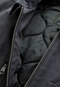 Mørkegrå jakke med en struktureret overflade og glat sort quilted foer. Har en robust lynlås og et label med mærkeoplysninger.