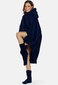 Navy kék kapucnis, oversized takaró poncsó, puha, bolyhos anyagból, bordázott mandzsettákkal és rövid, laza szabással. Kényelmes és otthonos dizájn.