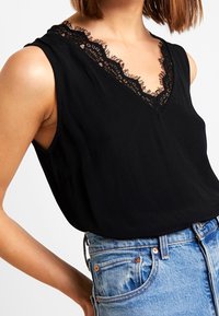 Haut sans manches noir avec un col en V, agrémenté d'une délicate bordure en dentelle le long du décolleté. Tissu lisse avec une coupe décontractée. Associé à un jean bleu clair.