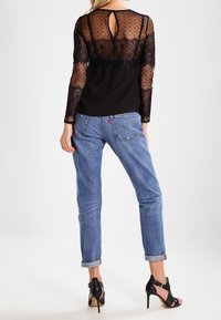 Schwarzes, durchsichtiges Langarmoberteil mit Polka-Dots, Spitzenakzenten und einem Schlüsselloch-Rückenausschnitt. Kombiniert mit blauen Jeans mit umgeschlagenem Saum und schwarzen High-Heel-Sandalen.