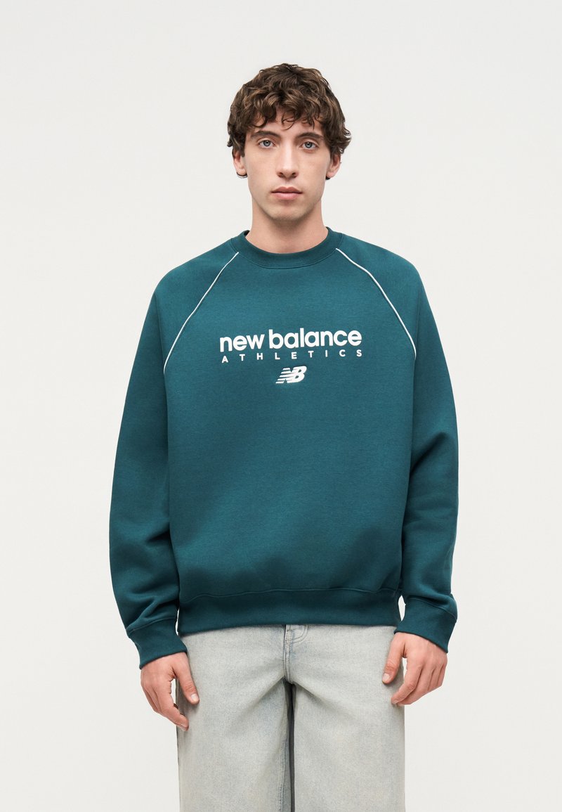 Jeune homme portant un sweat-shirt turquoise New Balance Athletics et un jean gris clair, debout devant un fond clair uni.