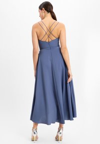 Langes, einfarbig blaufarbenes Kleid mit tailliertem Oberteil und gekreuzten Spaghettiträgern. Fließender Rock und verdeckter Reißverschluss hinten. Silberne Absatzsandalen.