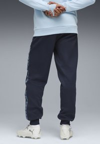 Marineblaue Sweatpants mit elastischen Bündchen, die einen gemusterten Seitenstreifen aufweisen. Kombiniert mit weißen Fußballschuhen und einem hellblauen Oberteil.