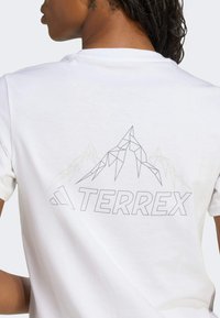Camiseta de algodón blanca con un gráfico geométrico de montaña y texto "TERREX" en gris en la parte posterior. Diseño de mangas cortas y cuello redondo.