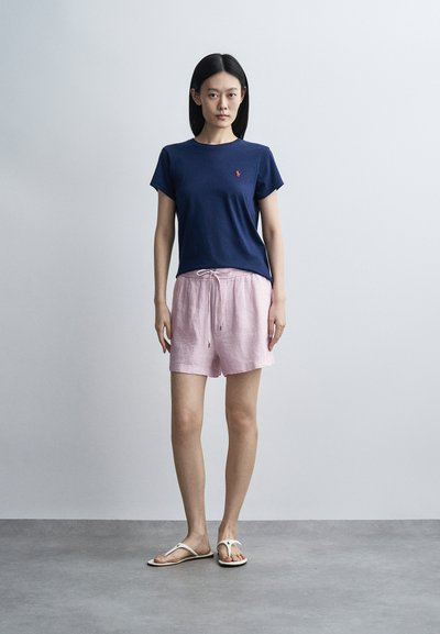 Γυναίκα με ίσια μαύρα μαλλιά που φοράει μπλε σκούρο μπλουζάκι, ανοιχτό ροζ σορτς με κορδόνι και λευκά σανδάλια, στέκεται μπροστά σε μια απλή γκρι φόντο.