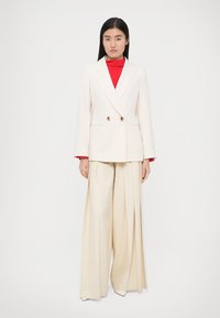 Beige double-breasted blazer met gouden knopen, gecombineerd met crèmekleurige, wijde broekspijpen. Rood coltruitje zichtbaar onder de blazer.