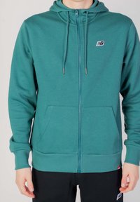 Tealfärgad zip-up hoodie med två framfickor, ribbad mudd och nederkant, justerbar dragsko på huven och en liten logotyp på vänster bröst.