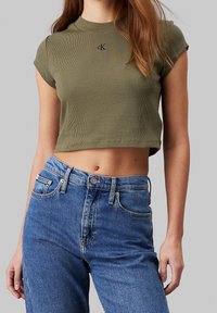 Olivově zelené zkrácené tričko z žebrované látky s kulatým výstřihem a krátkými rukávy, na hrudi s malým logem. Skombinováno s modrými džíny s vysokým pasem.
