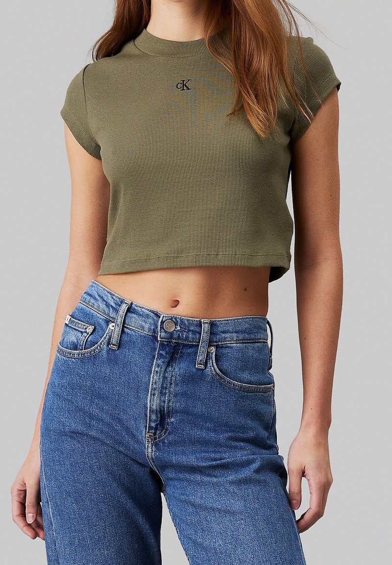 Olivově zelené zkrácené tričko z žebrované látky s kulatým výstřihem a krátkými rukávy, na hrudi s malým logem. Skombinováno s modrými džíny s vysokým pasem.