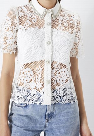 Weiße Spitzenbluse mit kurzen Ärmeln und Blumenmuster, perlenverzierten Knöpfen und einem mit Schmuck besetzten Kragen, kombiniert mit hellblauen Jeans.