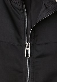 Gros plan sur une veste zippée noire, mettant en avant une texture de tissu lisse, une fermeture éclair en métal argenté et des détails de col côtelé. Design simple et fonctionnel.