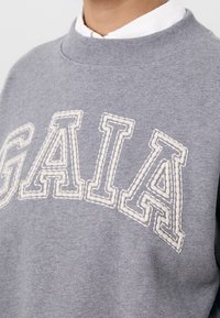 Grijze sweatshirt met gestructureerde witte stiksels die "GAIA" op de borst spellen. Klassiek ronde hals ontwerp met ribbenboord.