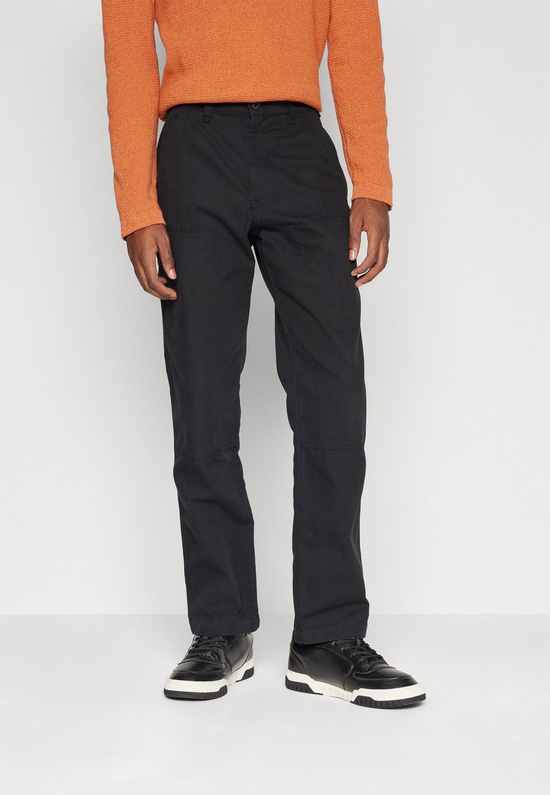Homme portant un pantalon noir droit, des baskets noires et blanches, et un pull orange à manches longues, debout devant un fond uni.