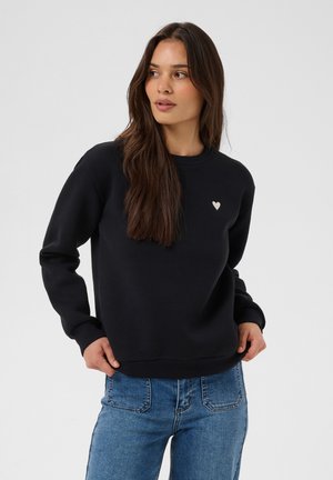 Femme aux longs cheveux bruns portant un sweat-shirt noir avec un petit cœur blanc sur la poitrine et un jean bleu, regardant sur le côté.