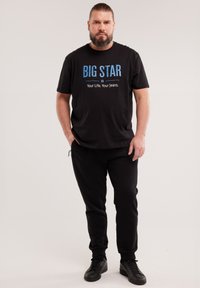 Habemega mees, kes kannab musta T-särki, millel on kiri "BIG STAR Your Life. Your Jeans.", musti pükse ja musti tossusid, seisab ühevärvilise tausta ees.