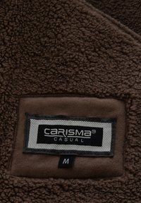 Bruine fleece stof met een prominente label waarop "CARISMA CASUAL" staat en maat "M" in witte tekst op een zwarte achtergrond. Zachte textuur.