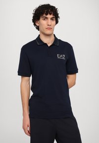 Tamsiai mėlynas polo marškinėliai, trumpomis rankovėmis, apykaklė su kontrastingu apvadu, kairėje krūtinės pusėje yra EA7 logotipas. Minkšta medžiaga, standartinė pritaikymo forma.