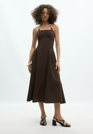 Femme aux cheveux bouclés portant une robe midi marron foncé sans manches avec un col halter et des talons noirs à brides sur un fond uni.