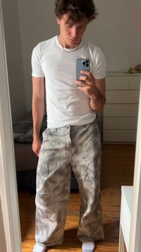 Giovane in maglietta bianca e pantaloni grigi tie-dye larghi, che si scatta un selfie allo specchio in una camera da letto con pavimento in legno.