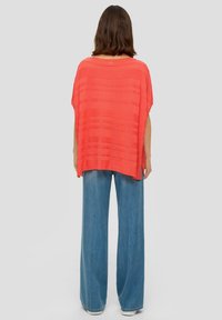 Koraalkleurige oversized top met gestructureerde strepen, korte mouwen en een brede silhouet, gecombineerd met lichtblauwe wijde jeans en witte schoenen.