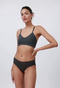 Ung kvinne med mørkt hår samlet bakover, iført en mørkegrå ribbestrikket bralette og matchende hipster-truser, stående med hånden på hoften.