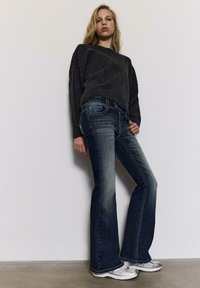 Grauer Strickpullover, dunkelblaue Schlagjeans mit leichten Ausbleichungen und silberne Sportschuhe. Glatte Stoffstrukturen und kontrastierende Nähte.