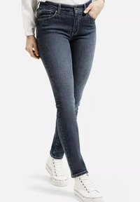 Kobieta w ciemnych dopasowanych jeansach i białych trampkach na wysokiej podeszwie, stojąca z jedną ręką w kieszeni na tle jednolitego tła.