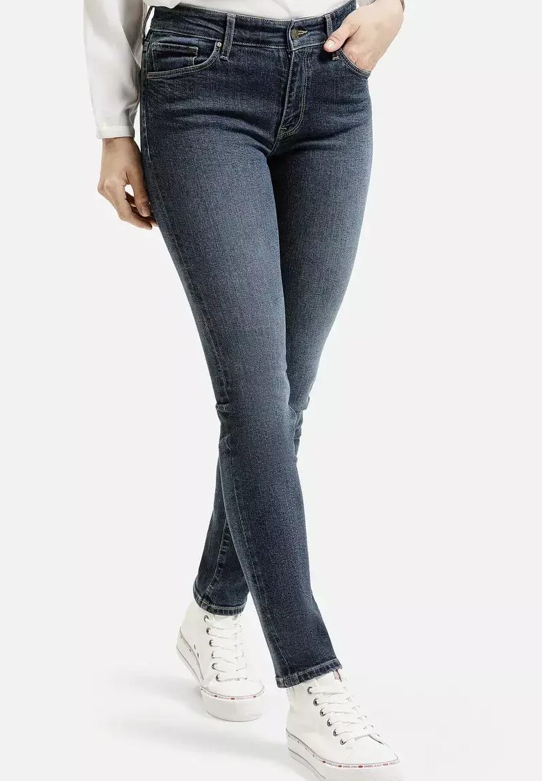 Kobieta w ciemnych dopasowanych jeansach i białych trampkach na wysokiej podeszwie, stojąca z jedną ręką w kieszeni na tle jednolitego tła.