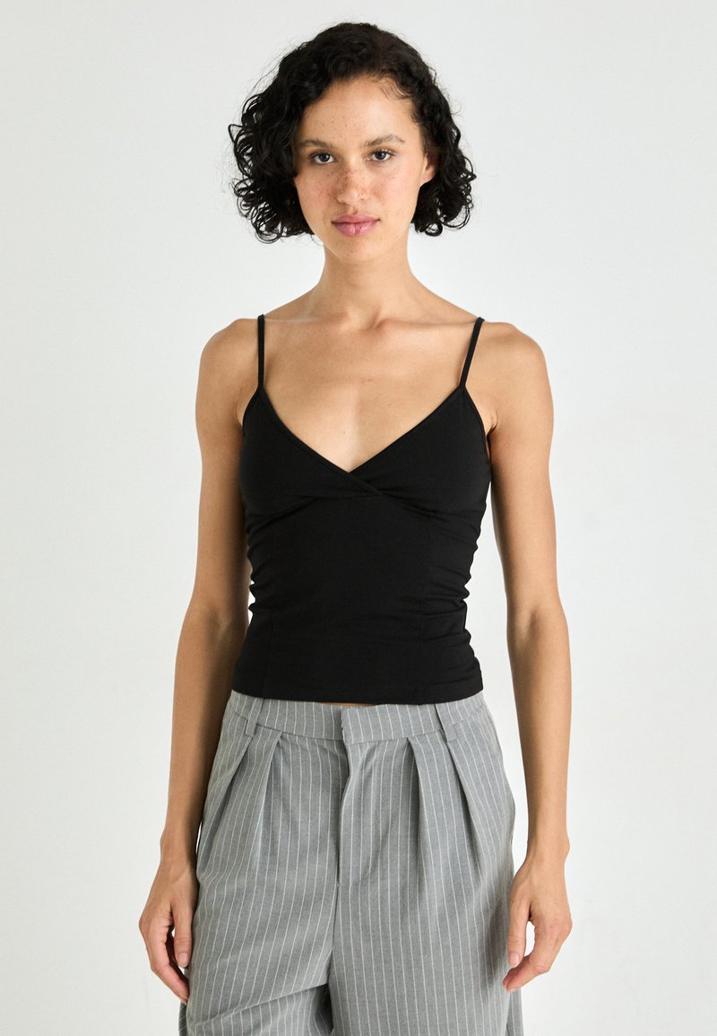 Black, fitted camisole with a V-neck and thin straps, made from a smooth, stretchy fabric. Worn with gray striped, pleated trousers.