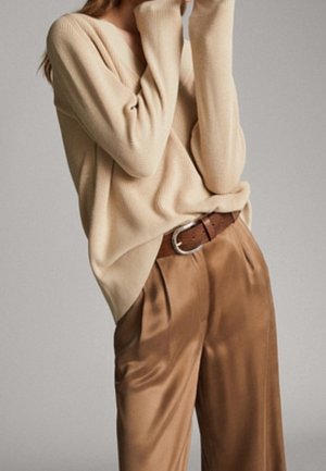 Femme portant un pull beige côtelé rentré dans un pantalon marron brillant taille haute avec une large ceinture en cuir marron.