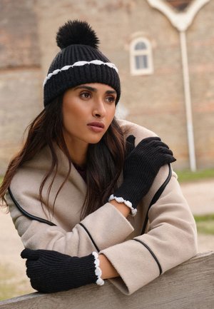 Mujer que lleva un abrigo beige con ribete negro, gorro de punto negro con pompón y guantes negros a juego con bordes blancos con volantes apoyándose en una barandilla de madera al aire libre.
