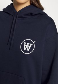Marinblå hoodies i mjukt tyg med en cirkulär vit logotyp med stiliserade bokstäver fram, snörning vid halsringningen.