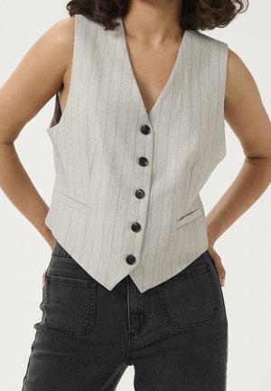 Kvinde iført en lys grå vest med tynde striber, knapper foran og frontlommer samt mørke højtaljede jeans med store forlommer.
