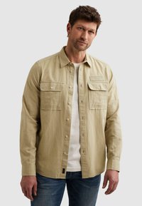 Homme portant une chemise beige à boutons avec deux poches poitrine par-dessus un t-shirt blanc et un jean bleu, debout devant un fond uni.