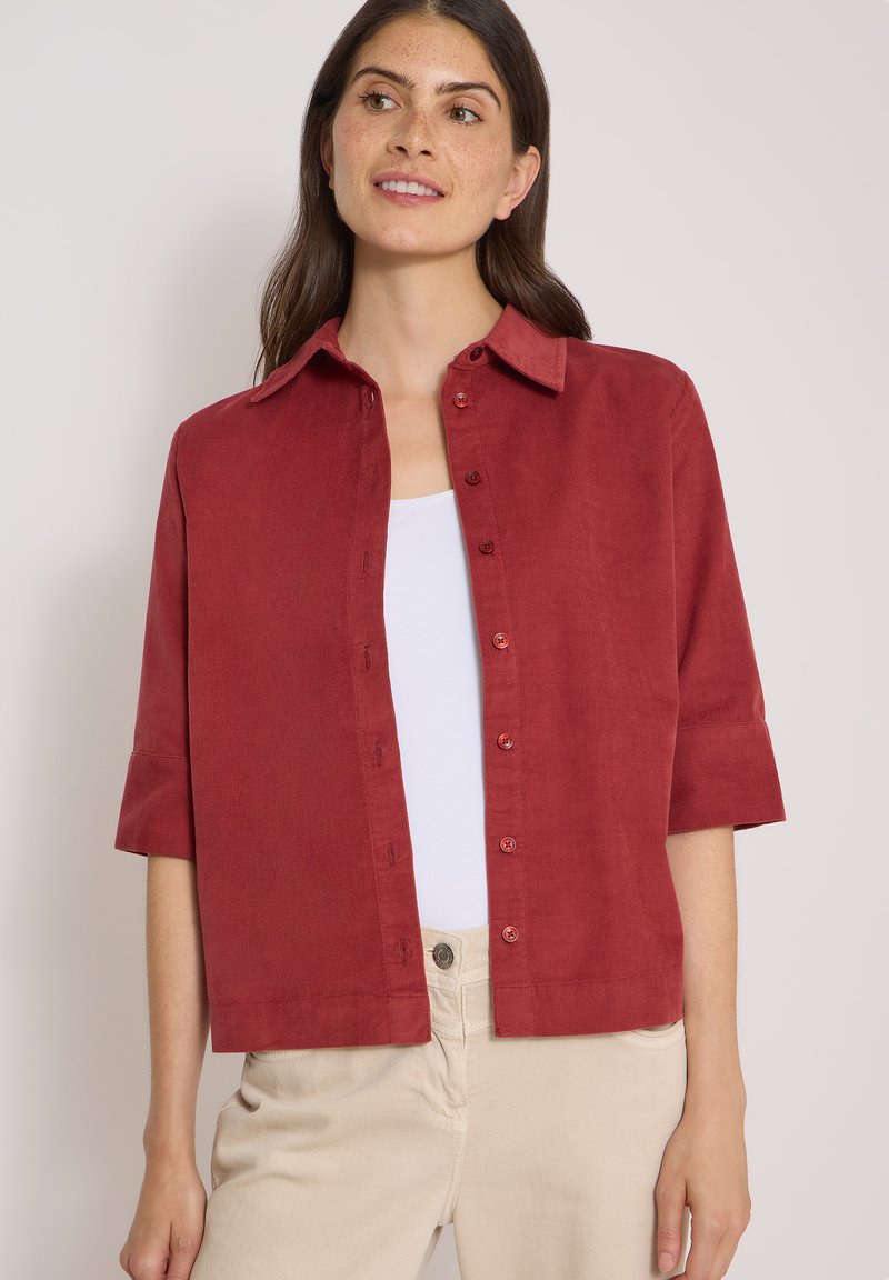Femme aux longs cheveux foncés portant une chemise rouge boutonnée par-dessus un haut blanc et un pantalon beige, debout devant un fond clair uni.