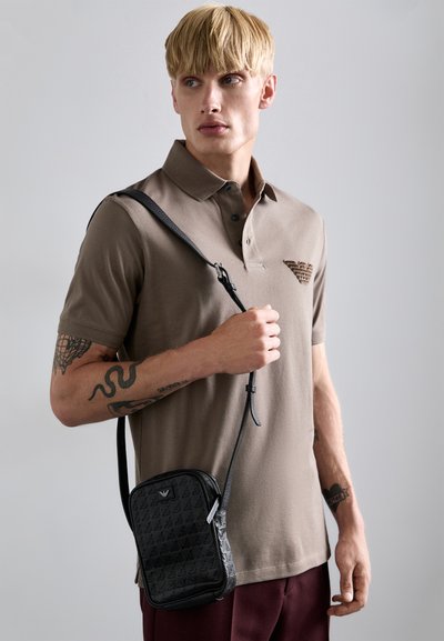 Sort crossbody taske med geometrisk mønster, lynlås lukning og justerbar rem; båret over en brun polo skjorte.