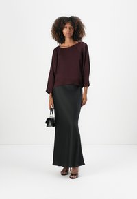 Haut bordeaux ample avec des manches larges associé à une jupe noire moulante et longue. Le mannequin tient un petit sac à main noir.