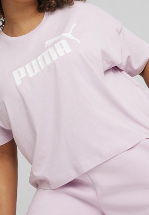 Osoba w jasnofioletowej koszulce Puma i pasujących spodniach, pokazująca górną część tułowia i część ramienia na jednolitym tle.