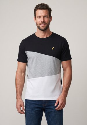 Homme portant un t-shirt bicolore noir, gris et blanc avec un petit logo d'oiseau jaune et un jean bleu, debout devant un fond uni.