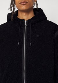 Schwarze Kapuzenjacke mit Reißverschluss aus plüschigem Material, ausgestattet mit einem glatten Reißverschluss, schwarzen Akzenten und einem kleinen Logo auf der linken Brustseite.