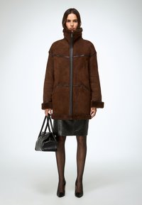 Chaqueta marrón de piel vuelta con cuello alto y cremallera, detalles en cuero negro, combinada con una falda negra texturizada y sosteniendo un bolso negro.