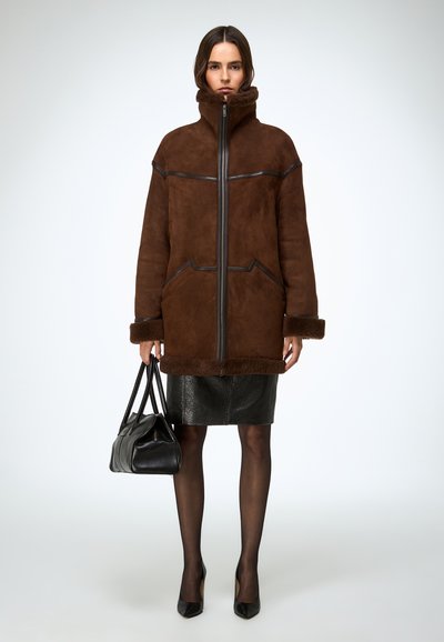 Bruine shearlingjas met hoge kraag en rits, zwarte leren accenten, gecombineerd met een getextureerde zwarte rok en een zwarte handtas in de hand.