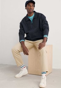 Pulover bleumarin cu buzunar frontal și accente turcoaz la manșete, asortat cu pantaloni bej și adidași albi cu glezna înaltă. Șosete cu dungi vizibile.