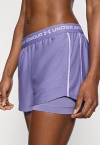 Μωβ αθλητικό σορτς κατασκευασμένο από ελαφρύ ύφασμα, με φαρδιά ζώνη μέσης που φέρε ι τη σήμανση "UNDER ARMOUR" και αντίθετες λεπτομέρειες στα πλαϊνά.