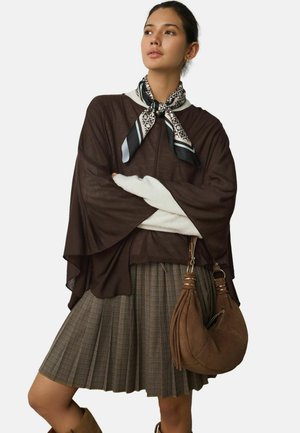 Frau trägt einen braunen Poncho, einen karierten Faltenrock, ein gemustertes Halstuch und trägt eine braune Wildleder-Umhängetasche.