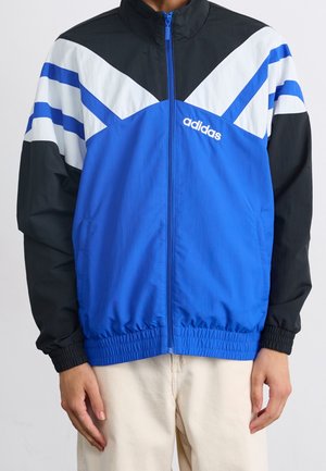 Homme portant une veste zippée Adidas bleu, noir et blanc avec des poignets et une ceinture élastiques, associée à un pantalon beige clair.