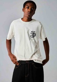 Camiseta de algodón blanca con mangas cortas y un diseño gráfico negro en el lado izquierdo del pecho; combinada con jeans de mezclilla oscura. Textura suave.