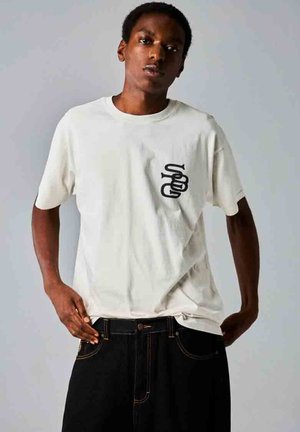 Camiseta de algodón blanca con mangas cortas y un diseño gráfico negro en el lado izquierdo del pecho; combinada con jeans de mezclilla oscura. Textura suave.