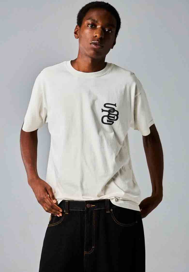 Camiseta de algodón blanca con mangas cortas y un diseño gráfico negro en el lado izquierdo del pecho; combinada con jeans de mezclilla oscura. Textura suave.