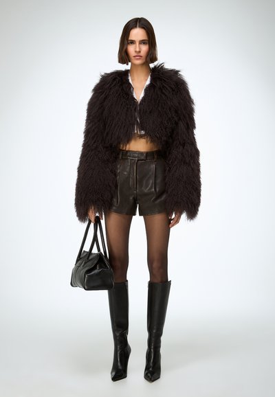 Zwarte jas van namaakbont, wit cropped shirt, bruine leren korte broek, zwarte kniehoge laarzen, met een zwarte handtas. Gladde en gestructureerde oppervlakken.